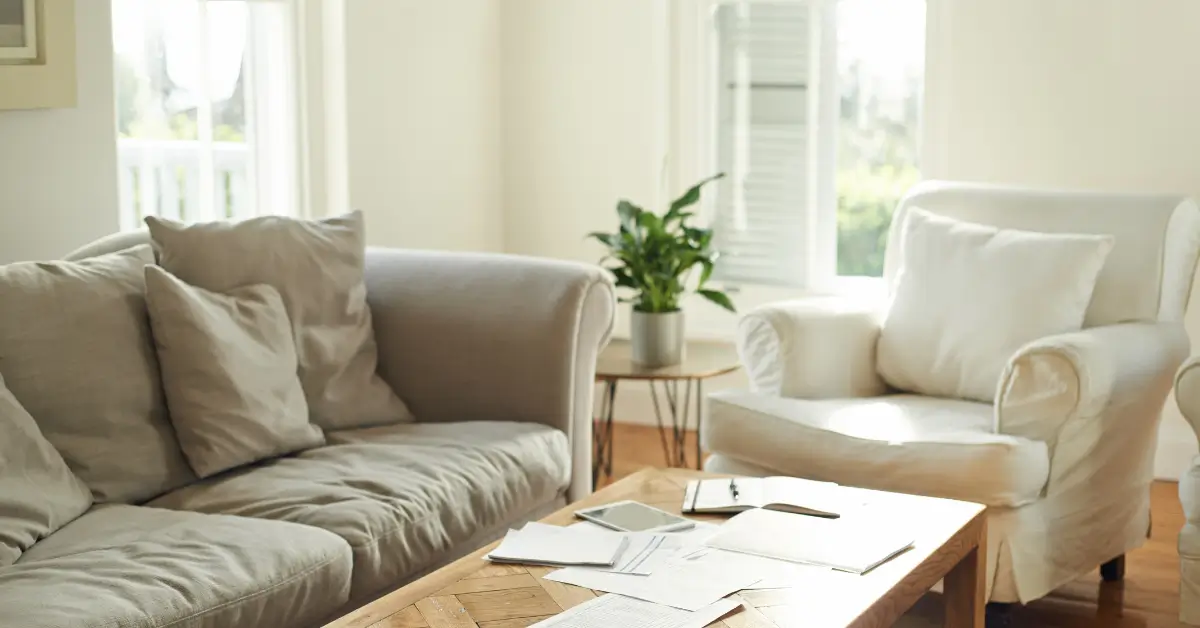 Bright living room with a beige sofa, white armchair, coffee table covered with papers, and a potted plant near sunlit windows.
